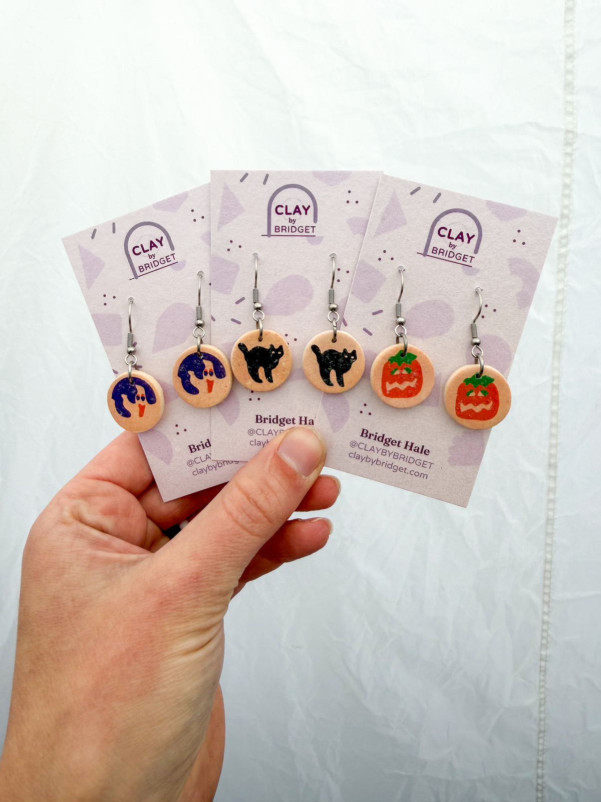 Three pairs of earrings held in a hand against a white background. They feature circular earrings that resemble Halloween cookies. From left to right, the designs are ghost, cat, and jack-o-lantern. 