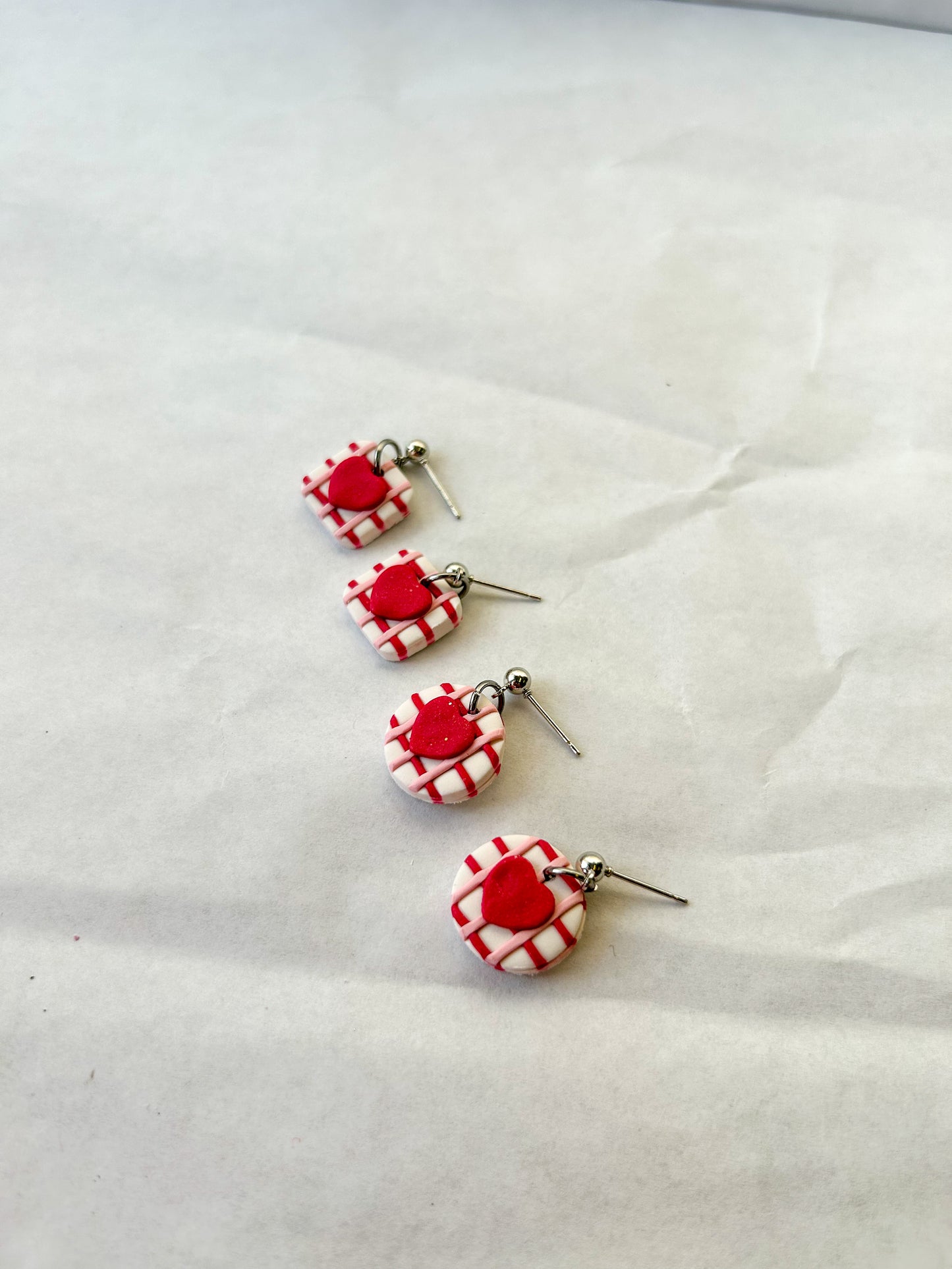 Two pairs of earrings laying next to each other on a flat background. One pair is square with hearts in the center and one pair is circle with hearts in the center