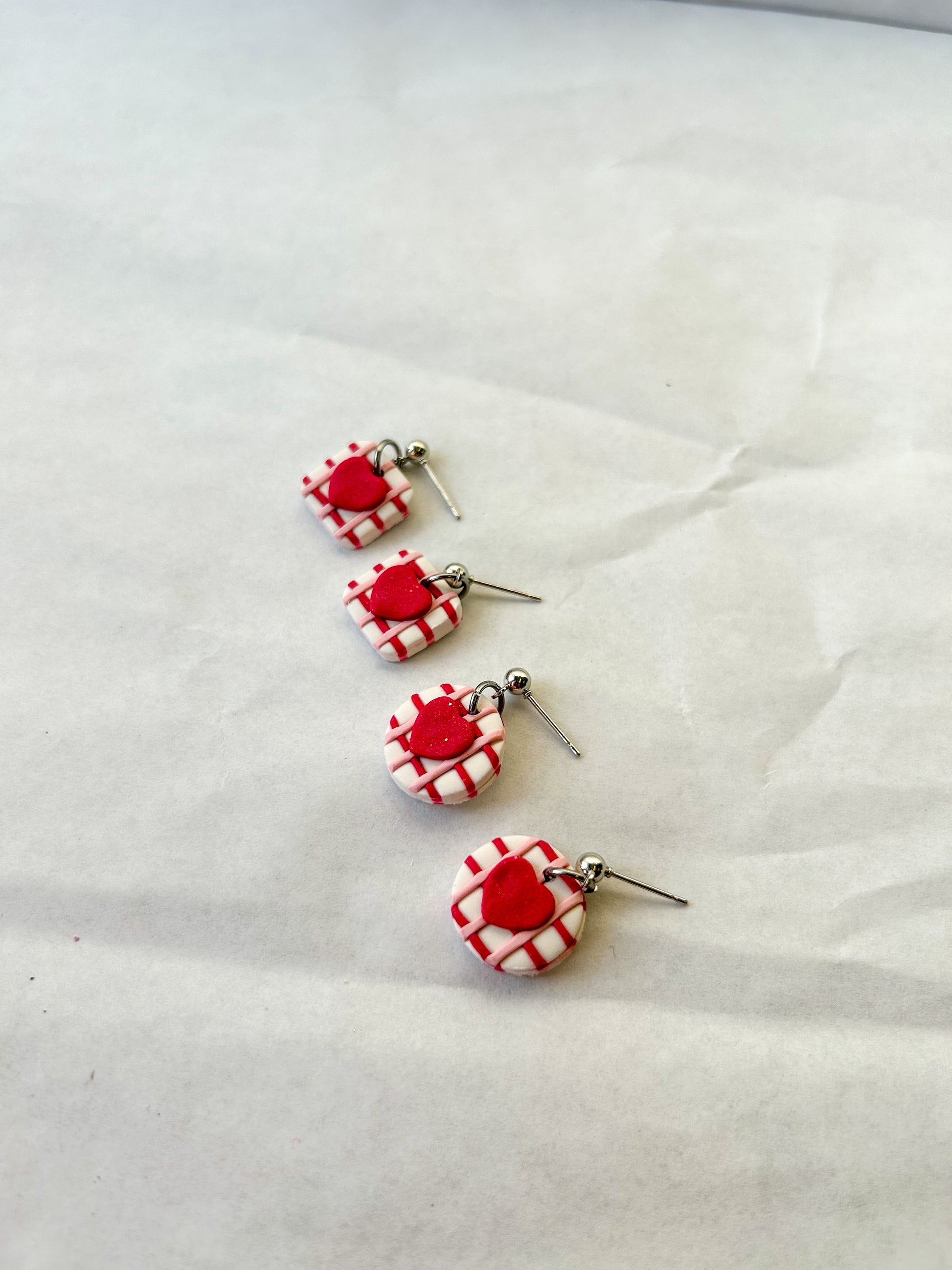 Two pairs of earrings laying next to each other on a flat background. One pair is square with hearts in the center and one pair is circle with hearts in the center