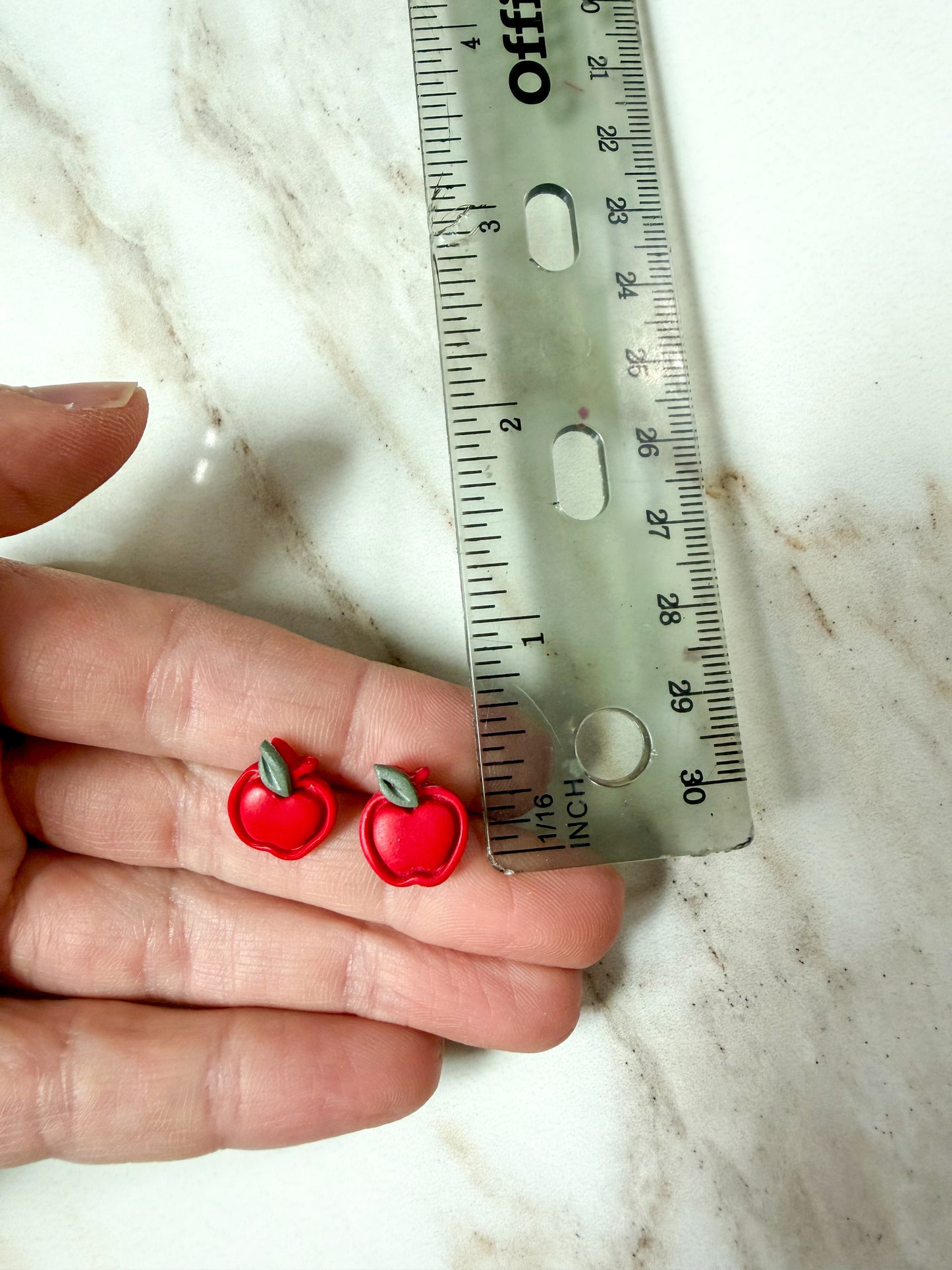 A close-up of a hand holding two small red apple-shaped earrings next to a ruler on a light marble surface. About 1/2 inch long. 