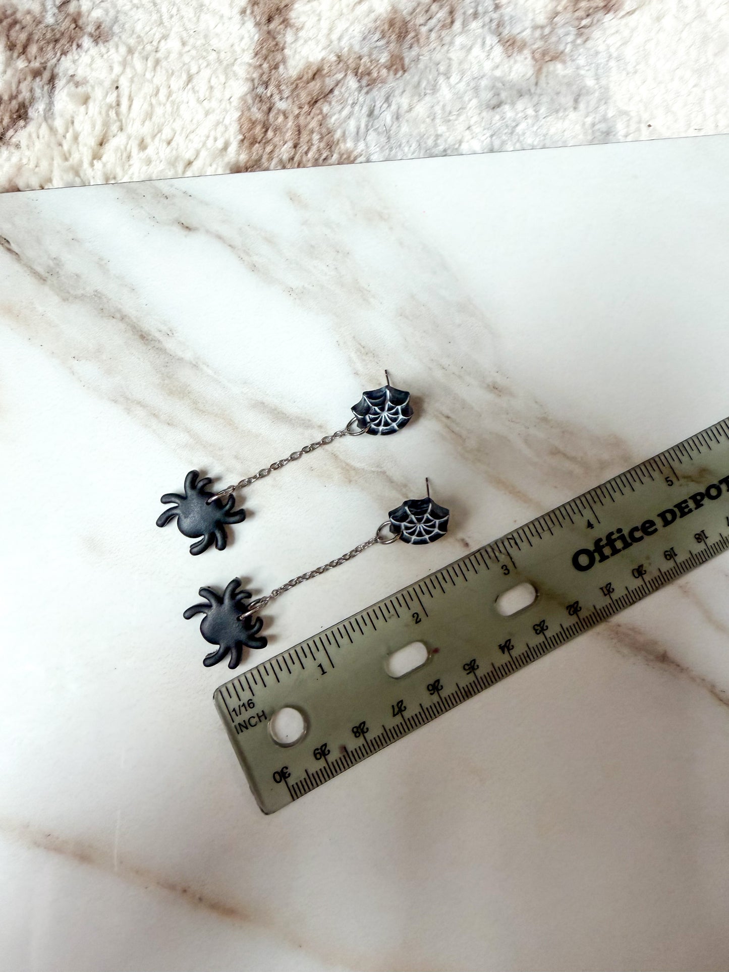 A pair of black spider drop earrings with spiderweb charms, displayed on a white marble surface next to a ruler for scale. Around 3 inches in length. 