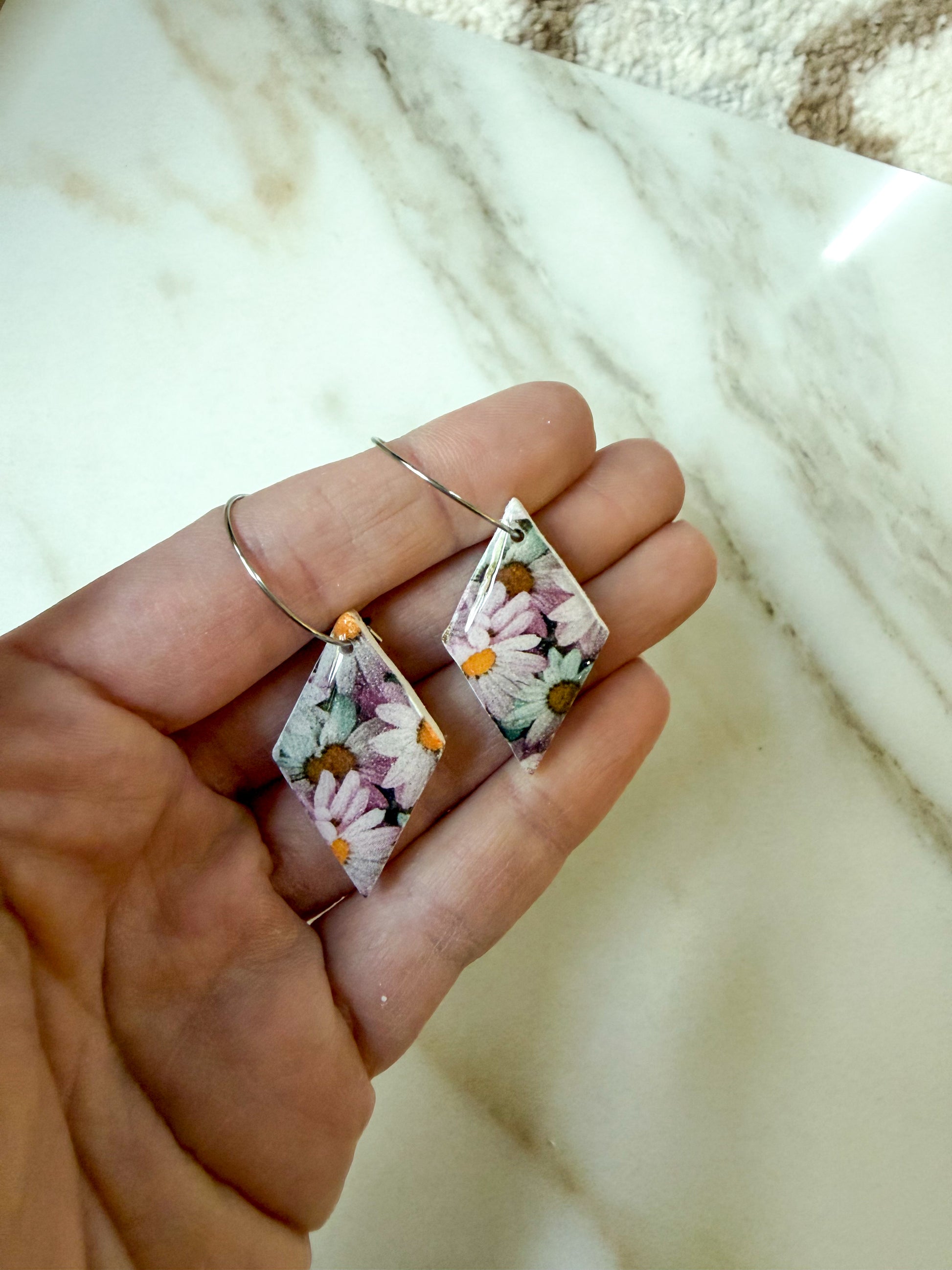 A hand holding a pair of dangling earrings with floral-patterned diamond-shaped charms and thin metal wire hoops, set against a light marble surface.