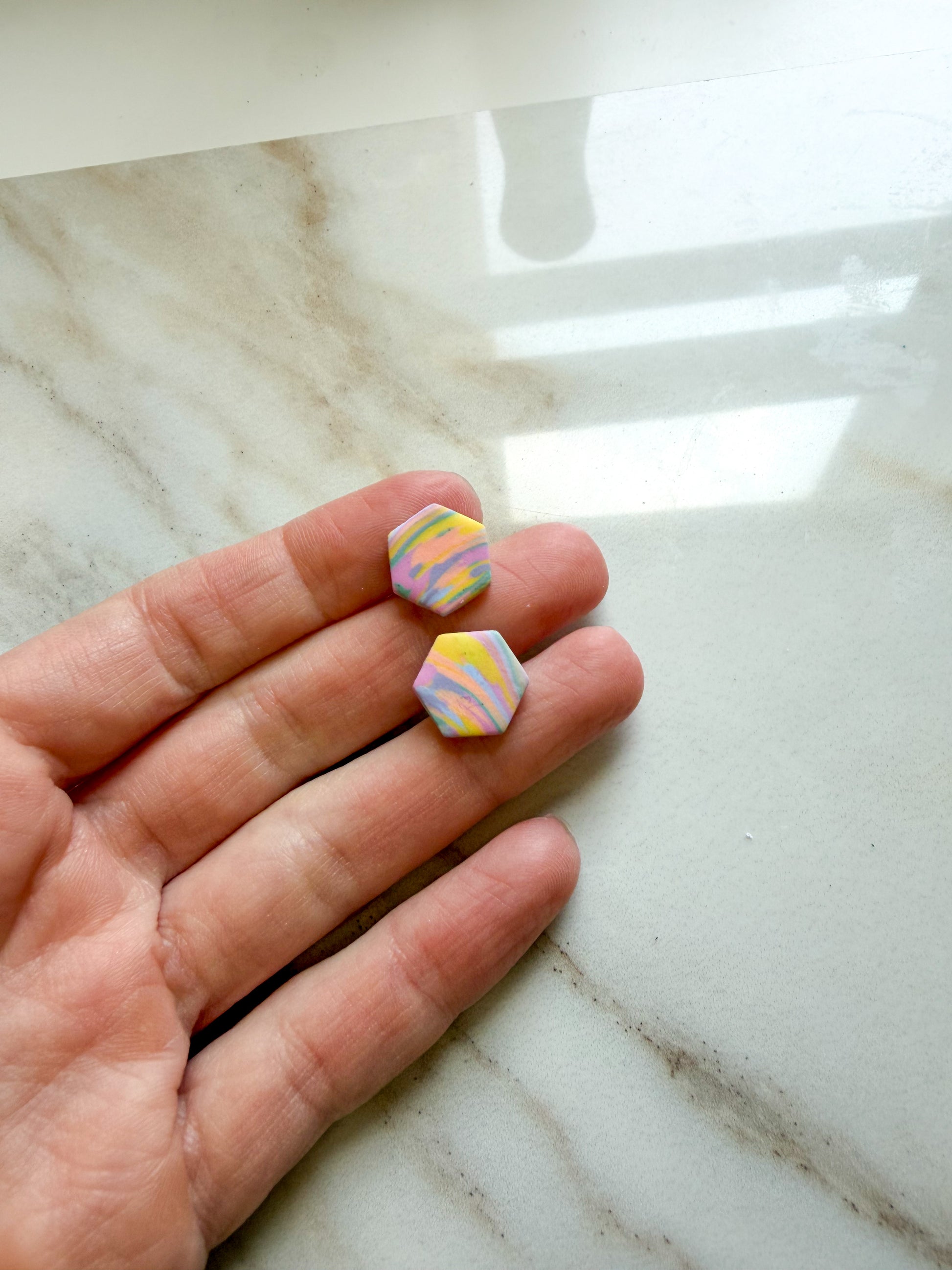 Two small hexagonal stud earrings with a pastel marbled pattern of pink, yellow, and blue, held on a person's fingers over a light marble surface.