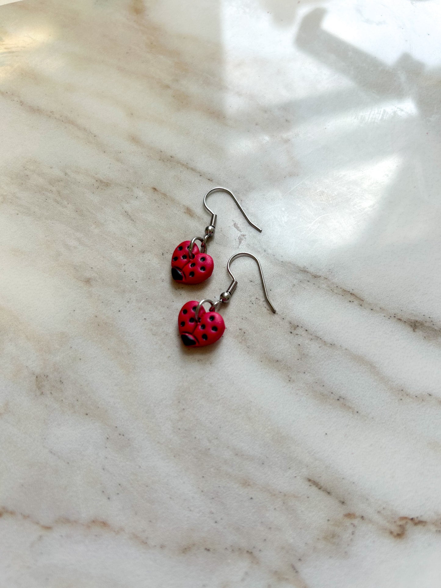 Heart shaped earrings that are painted to look like ladybugs, playing on a stone surface ￼