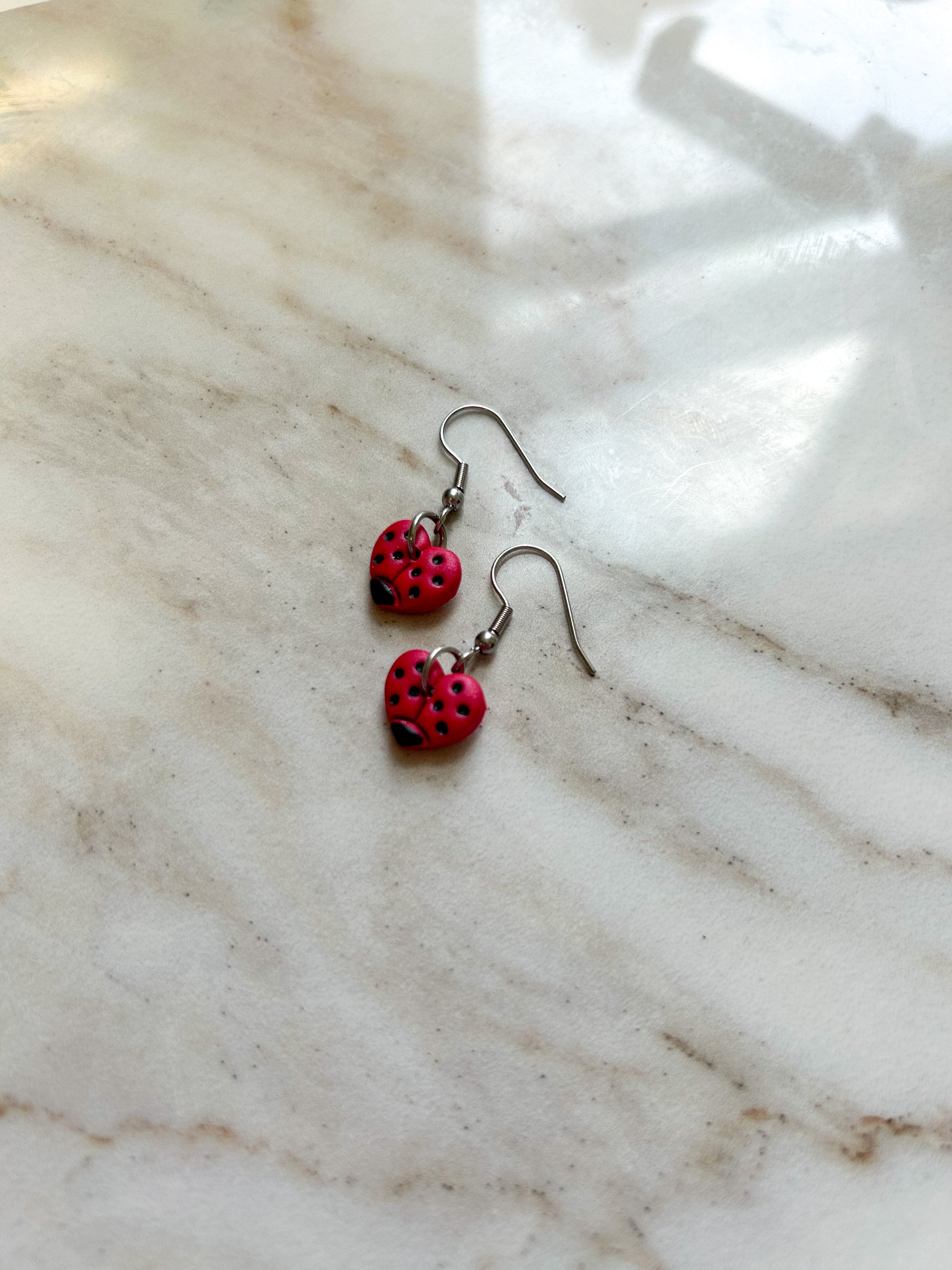 Heart shaped earrings that are painted to look like ladybugs, playing on a stone surface ￼