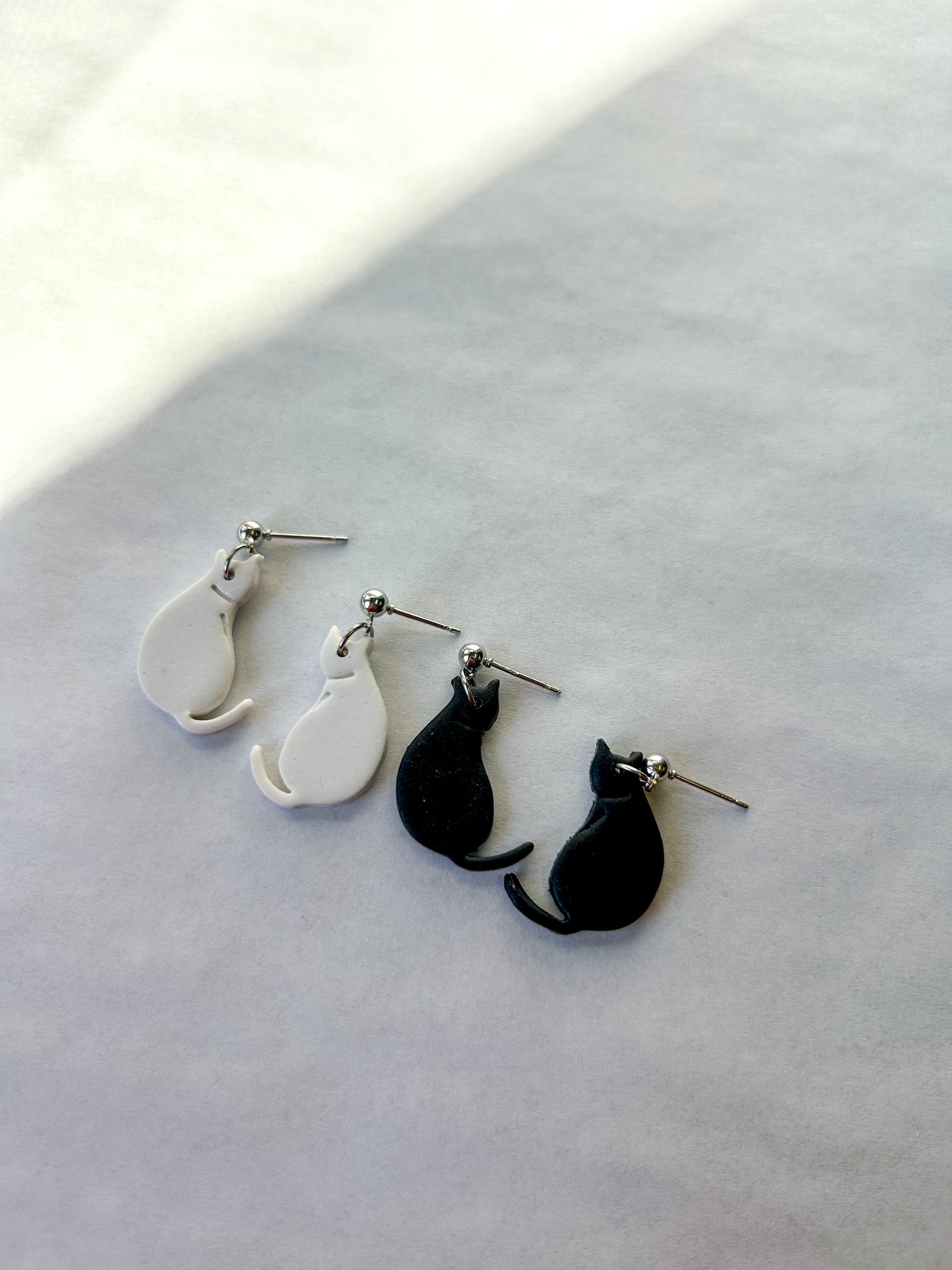 Two pairs of cat-shaped earrings on a light gray surface. One pair is white and one is black. 