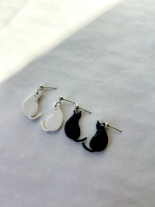 Two pairs of cat-shaped earrings on a light gray surface. One pair is white and one is black. 