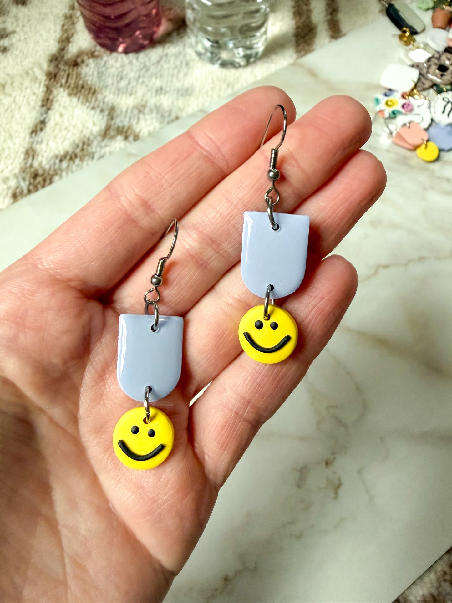 Hand holding a pair of earrings with smiley face charms on a marble surface.
