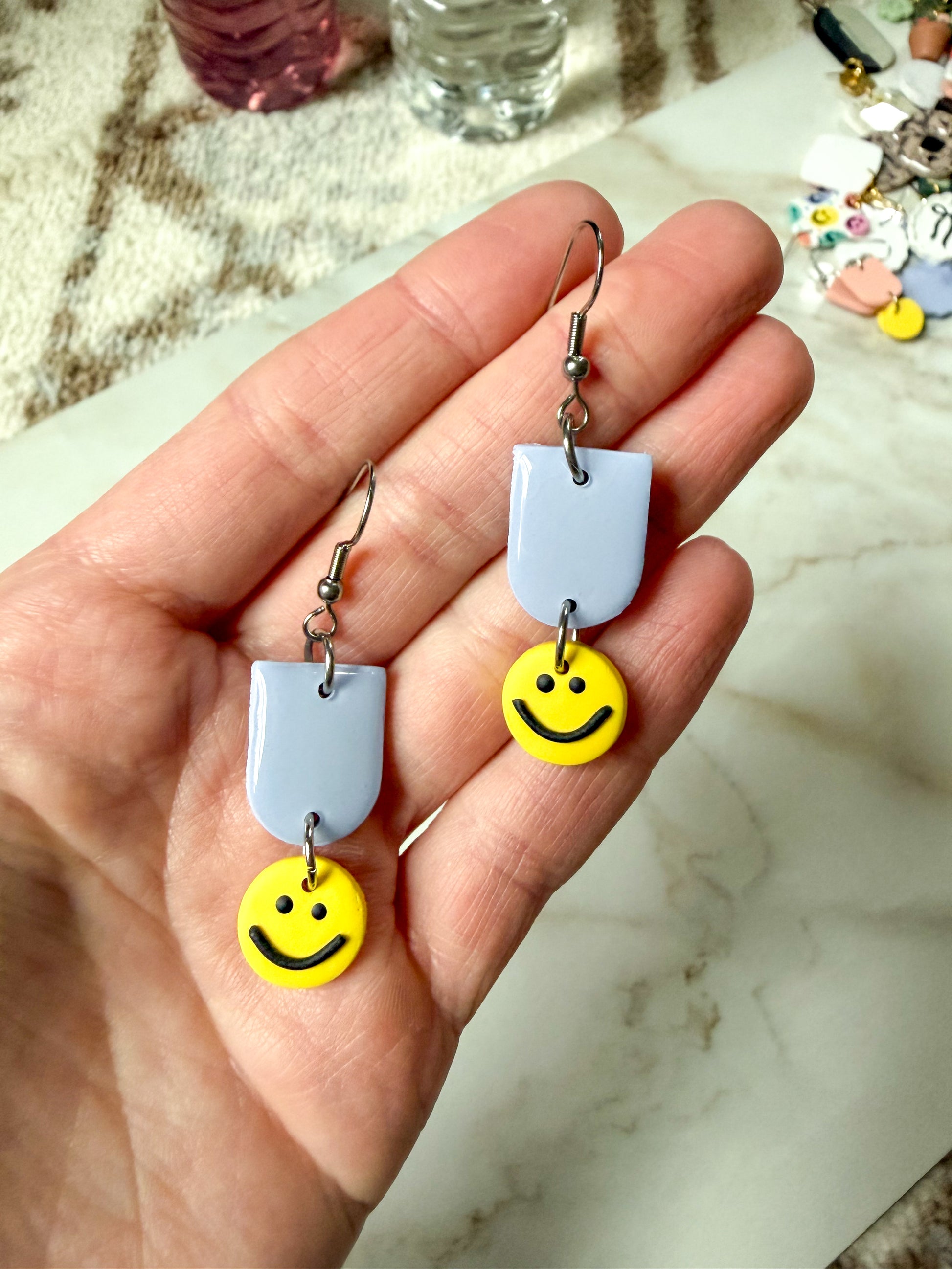 Hand holding a pair of earrings with smiley face charms on a marble surface.